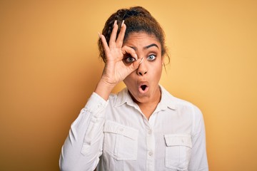 Young beautiful african american girl wearing casual shirt standing over yellow background doing ok gesture shocked with surprised face, eye looking through fingers. Unbelieving expression.