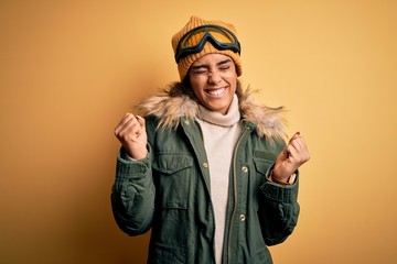 Young african american afro skier girl wearing snow sportswear and ski goggles very happy and excited doing winner gesture with arms raised, smiling and screaming for success. Celebration concept.