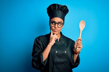 Naklejka premium Young african american chef girl wearing cooker uniform and hat holding wooden spoon tool serious face thinking about question, very confused idea