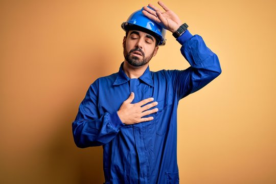 Mechanic man with beard wearing blue uniform and safety helmet over yellow background Touching forehead for illness and fever, flu and cold, virus sick