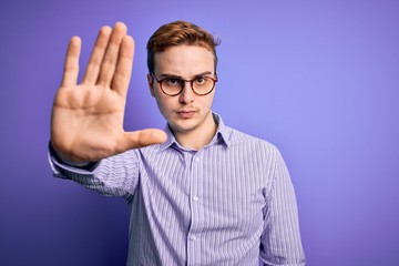 Naklejka premium Young handsome redhead man wearing casual shirt and glasses over purple background doing stop sing with palm of the hand. Warning expression with negative and serious gesture on the face.