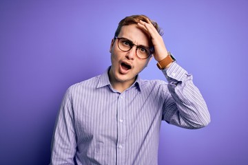 Young handsome redhead man wearing casual shirt and glasses over purple background surprised with hand on head for mistake, remember error. Forgot, bad memory concept.