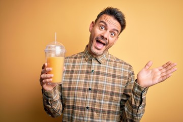 Young handsome man drinking healthy orange juice using straw over yellow background very happy and excited, winner expression celebrating victory screaming with big smile and raised hands