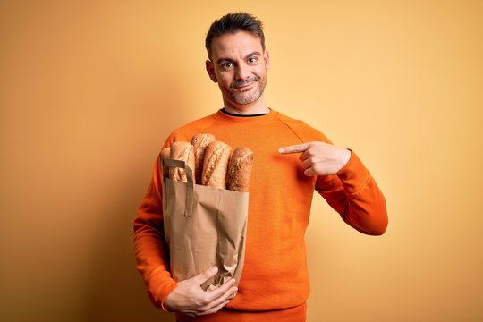 Young handsome man holding paper bag with bread over isolated yellow background with surprise face pointing finger to himself