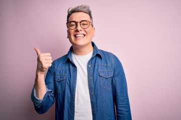 Young handsome modern man wearing glasses and denim jacket over pink isolated background smiling with happy face looking and pointing to the side with thumb up.