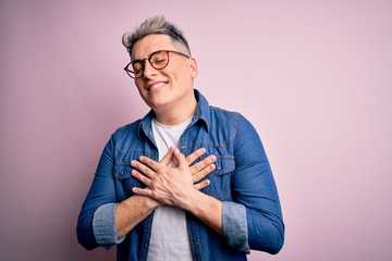 Young handsome modern man wearing glasses and denim jacket over pink isolated background smiling with hands on chest with closed eyes and grateful gesture on face. Health concept.
