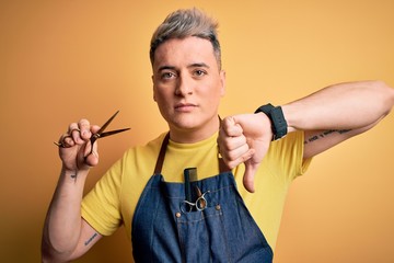 Young modern professional hairdresser man holding scissors over isolated yellow background with angry face, negative sign showing dislike with thumbs down, rejection concept