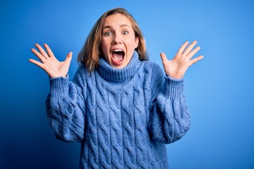 Young beautiful blonde woman wearing casual turtleneck sweater over blue background celebrating crazy and amazed for success with arms raised and open eyes screaming excited. Winner concept
