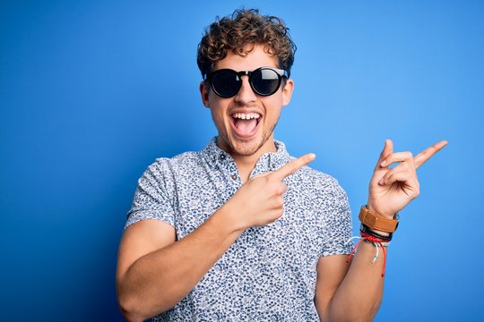 Young blond handsome man with curly hair on vacation wearing striped polo and sunglasses smiling and looking at the camera pointing with two hands and fingers to the side.