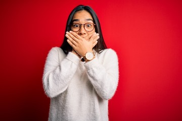 Young beautiful asian woman wearing casual sweater and glasses over red background shocked covering mouth with hands for mistake. Secret concept.