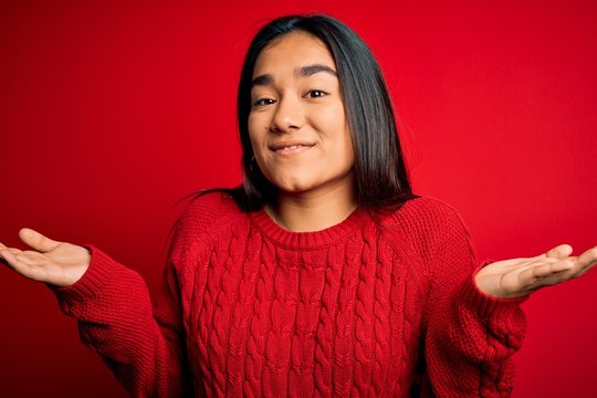 Young beautiful asian woman wearing casual sweater standing over isolated red background clueless and confused expression with arms and hands raised. Doubt concept.