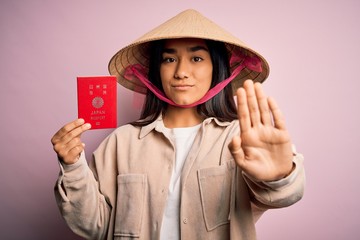 Young thai woman wearing traditional conical asian hat holding japan japanese passport with open hand doing stop sign with serious and confident expression, defense gesture