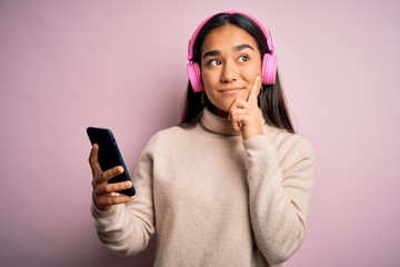 Young beautiful asian woman listening to music using smartphone and headphones serious face thinking about question, very confused idea