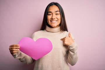 Young beautiful asian woman holding pink heart standing over isolated background with surprise face pointing finger to himself