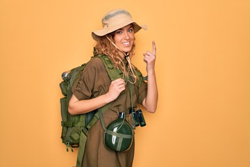 Young blonde explorer woman with blue eyes hiking wearing backpack and water canteen pointing finger up with successful idea. Exited and happy. Number one.