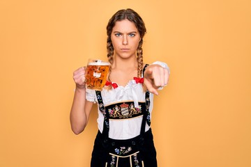 Beautiful blonde german woman with blue eyes wearing octoberfest dress drinking jar of beer pointing with finger to the camera and to you, hand sign, positive and confident gesture from the front