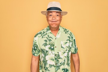 Middle age senior grey-haired man wearing summer hat and floral shirt on beach vacation puffing cheeks with funny face. Mouth inflated with air, crazy expression.
