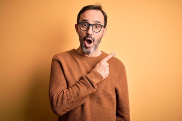 Middle age hoary man wearing brown sweater and glasses over isolated yellow background Surprised...
