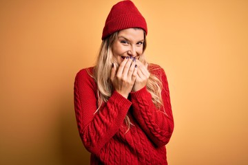 Young beautiful blonde woman wearing casual sweater and wool cap over white background laughing and embarrassed giggle covering mouth with hands, gossip and scandal concept