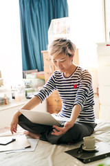 Beautiful Asian woman sitting on bed & working from home on laptop