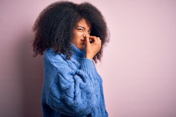 Young beautiful african american woman with afro hair wearing winter sweater over pink background smelling something stinky and disgusting, intolerable smell, holding breath with fingers on nose. 