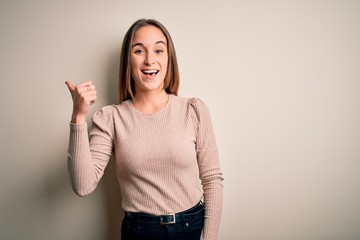 Young beautiful woman wearing casual sweater standing over isolated white background smiling with happy face looking and pointing to the side with thumb up.