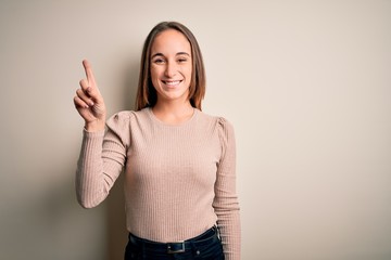 Young beautiful woman wearing casual sweater standing over isolated white background showing and pointing up with finger number one while smiling confident and happy.