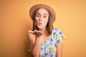 Young beautiful woman wearing casual t-shirt and summer hat over isolated yellow background looking at the camera blowing a kiss with hand on air being lovely and sexy. Love expression.