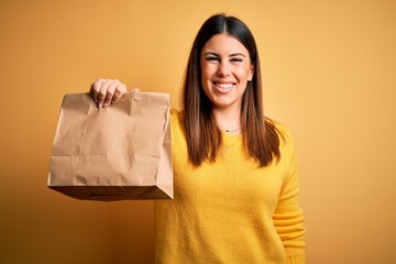 Young beautiful woman holding take away paper bag from delivery over yellow background with a happy face standing and smiling with a confident smile showing teeth