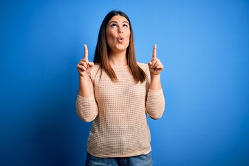 Young beautiful woman wearing casual sweater over blue background amazed and surprised looking up and pointing with fingers and raised arms.