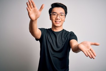 Young handsome chinese man wearing black t-shirt and glasses over white background looking at the camera smiling with open arms for hug. Cheerful expression embracing happiness.