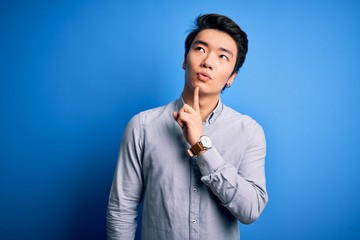 Young handsome chinese man wearing casual shirt standing over isolated blue background Thinking concentrated about doubt with finger on chin and looking up wondering