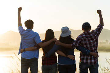 Group of happy friends are having fun with raised arms together in front of mountain and enjoy sunrise sunset showing unity and teamwork. Friendship happiness leisure partnership team concept.