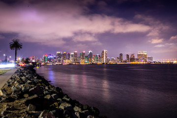 The lights of San Diego's skyline reflect off the waters of the Pacific Ocean, with sailboats in the foreground.