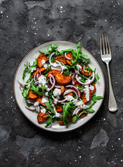 Baked crispy sweet potatoes, arugula, red onion and yogurt mustard dressing salad on a dark background, top view