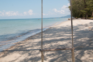 A wooden swing is hanging next to a clean tropical beach with blue sky background