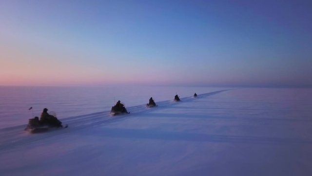 Unique flight close with snowmobiles fast speed ride Arctic extreme. Group of tourists no face in helmets. Clear sky sunny sunset horizon. Aerial drone footage.
