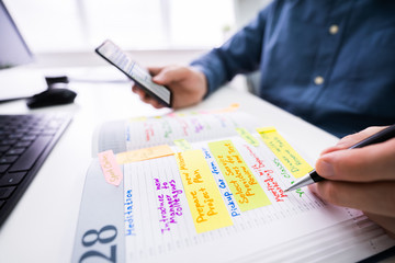 Businessman Holding Cellphone Writing Schedule In Diary With Pen