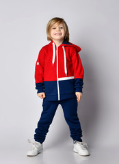 Full growth portrait of happy smiling blond kid boy in red and blue sportwear and white sneakers