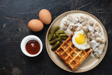 Beige tray with belgian potato waffle, champignons, fried egg and pickles. Top view on a dark brown stone background with space, studio shot