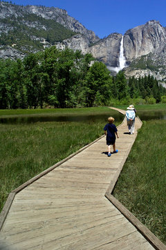 Grandson following grandparents crossing Sentinel Meadow on wood-plank path with Upper Yosemite Falls in background