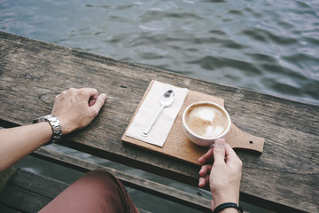 Sitting by a lake  and hand holding a cup of coffee with latte art serving by wooden tray.
