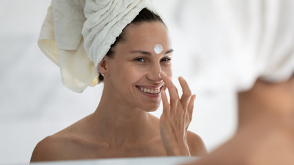 Woman reflected in mirror after morning shower with towel on head applies day facial cream close...