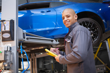 Charming male car service worker smiling to the camera while taking notes on his clipboard, lifted automobile on background