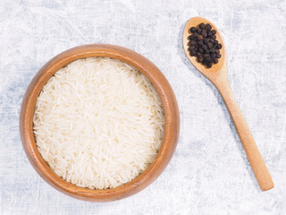 Indian long grain Basmati rice in brown wooden bowl and black pepper in spoon on white concrete background