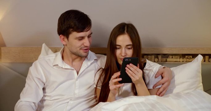 A young love couple: a Girl and a guy in white clothes are lying in a light white bed, looking at the phone, smiling, hugging and kissing, they are in a room.