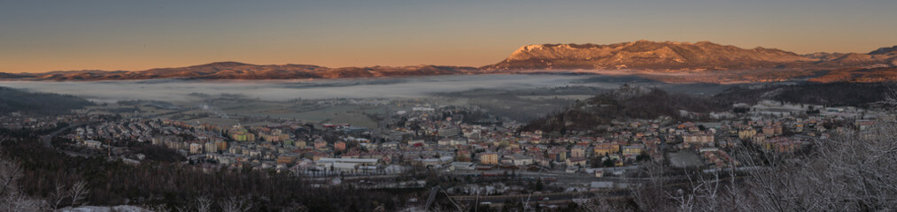 Fototapeta na wymiar Panorama of the city of Postojna in Slovenia in early winter morning with sun just rising up and lighting mountain ridge of Nanos in the background. Beautiful early panorama of a small city.
