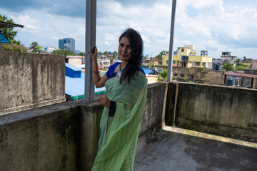 Portrait of a beautiful smiling brunette Indian Bengali woman in traditional wear sari standing on the roof top in sunny morning of Durga Puja festival in green natural background. Indian lifestyle.
