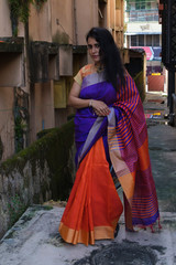 Portrait of a beautiful brunette Indian bengali thoughtful woman in traditional wear sari standing in a narrow lane in the morning of Durga Puja festival in urban background. Indian lifestyle.