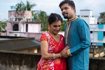 Naklejka premium A brunette Indian Bengali romantic couple in traditional wear enjoying themselves standing on the roof top in sunny morning of Durga Puja festival in green natural background. Indian lifestyle.